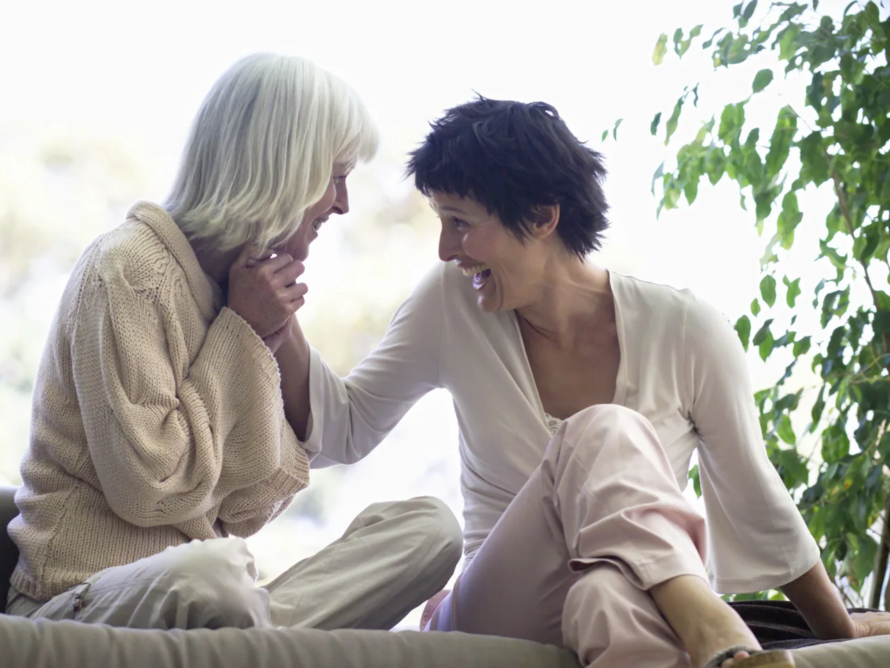 Aydınlık bir odada yan yana oturan iki kadın birbirine bakıp gülüyor.